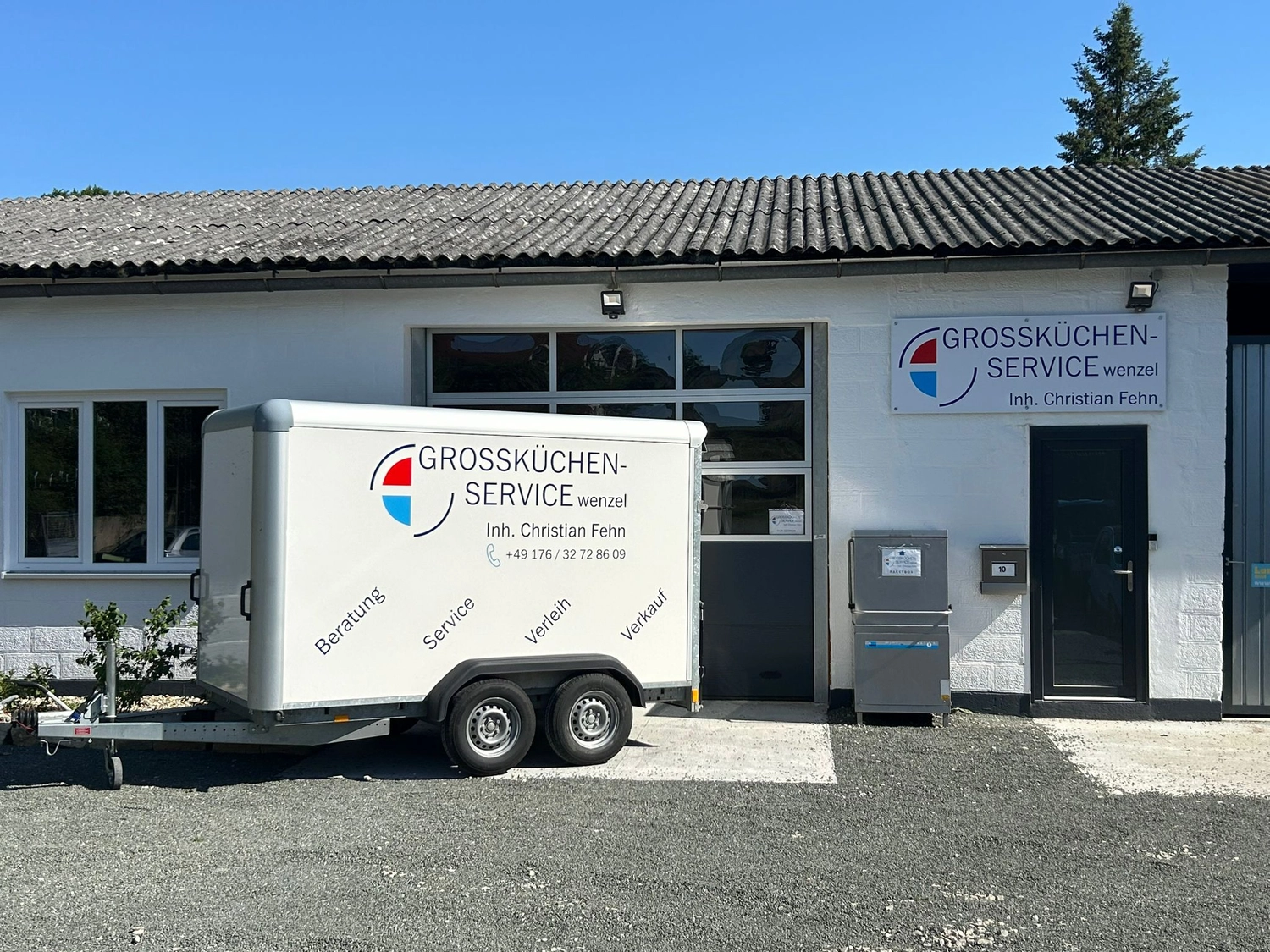Großküchenservice Wenzel - Großküchen Service in Forchheim - Auf einem Schotterplatz unter strahlend blauem Himmel stehen ein weißer Anhänger und ein Gebäude der Firma "GROSSKÜCHEN-SERVICE wenzel".